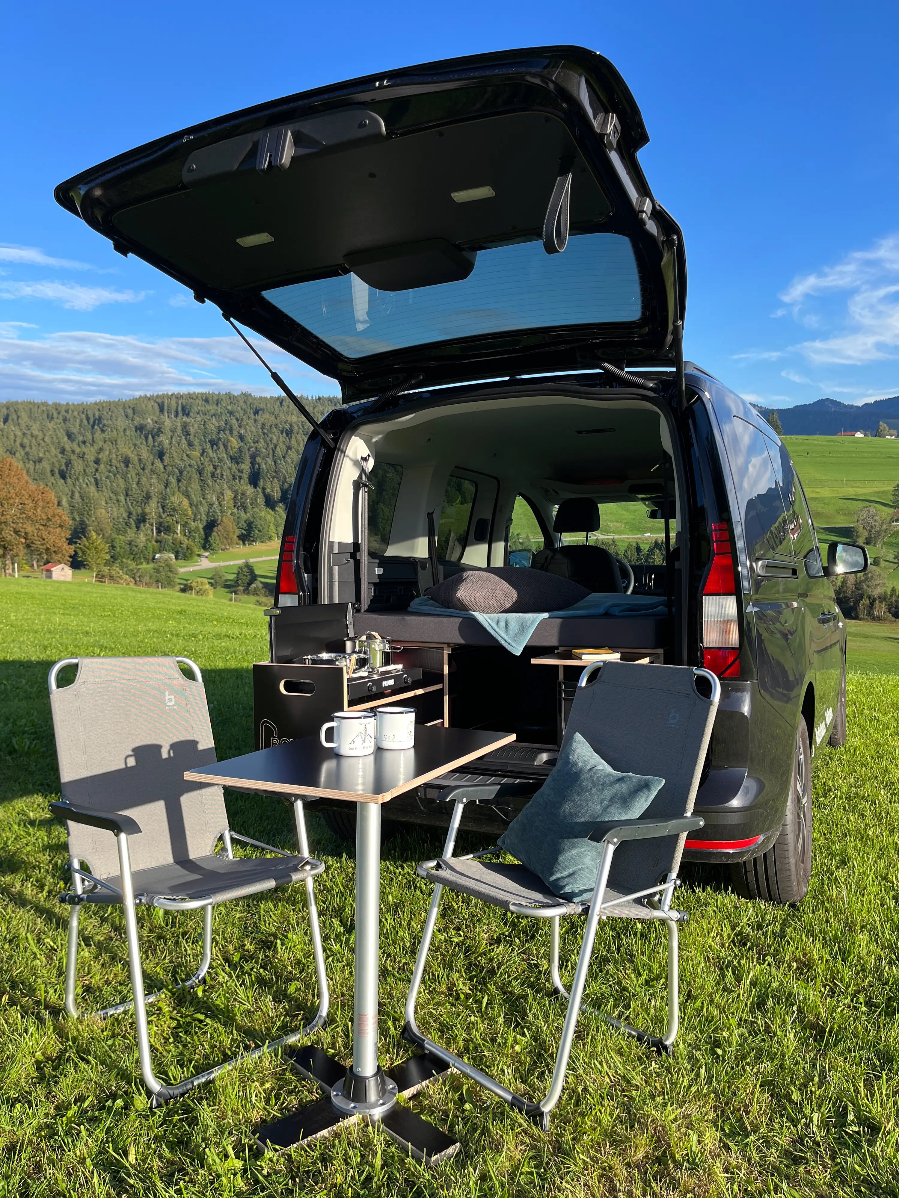 Van mit offener Heckklappe, Tisch und Stühlen, aufgestellt im Freien auf einer Wiese mit Bergen im Hintergrund.